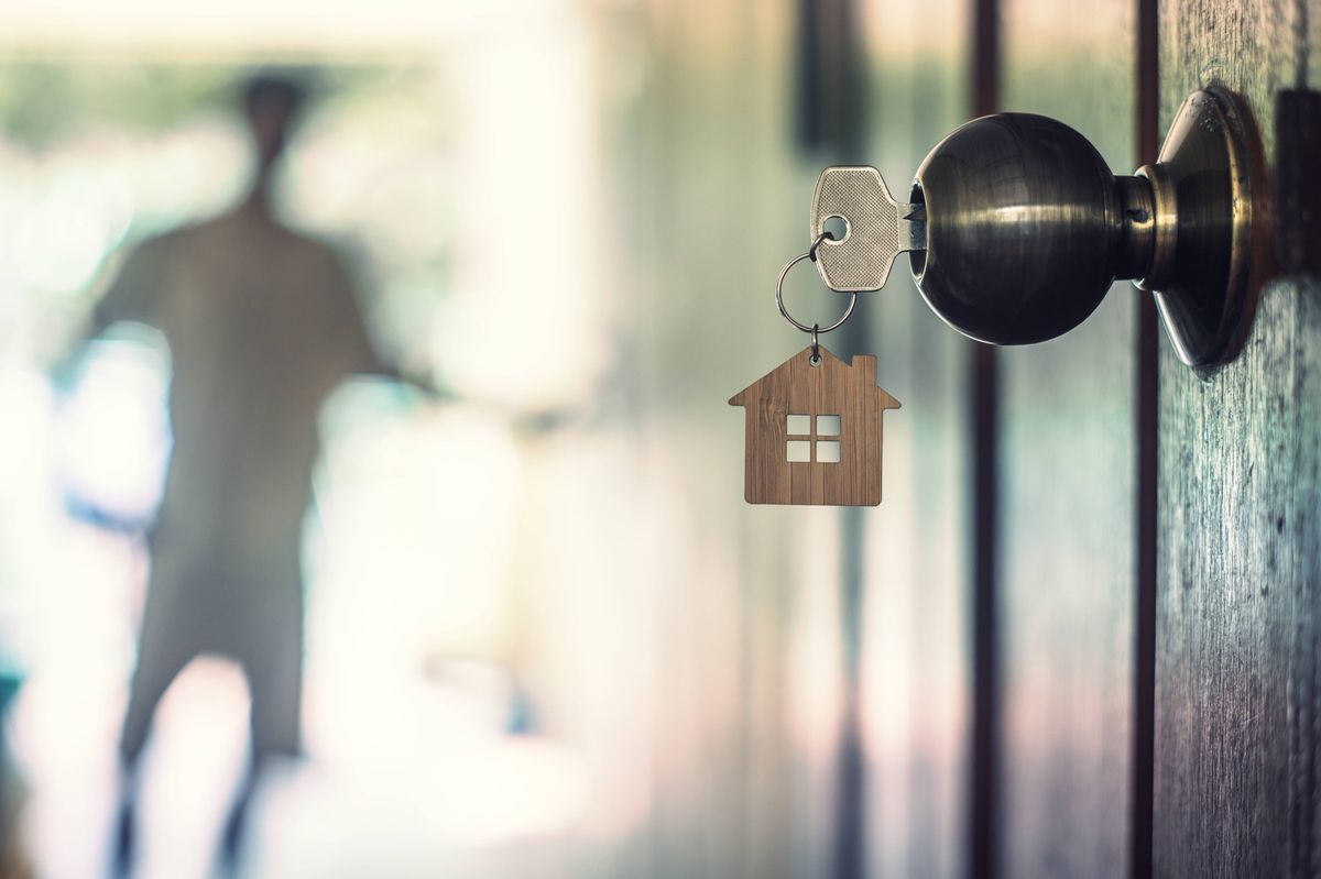 A key with a miniature house keychain is attached to a door handle, while a blurred figure is visible in the background, perhaps entering or exiting a room.