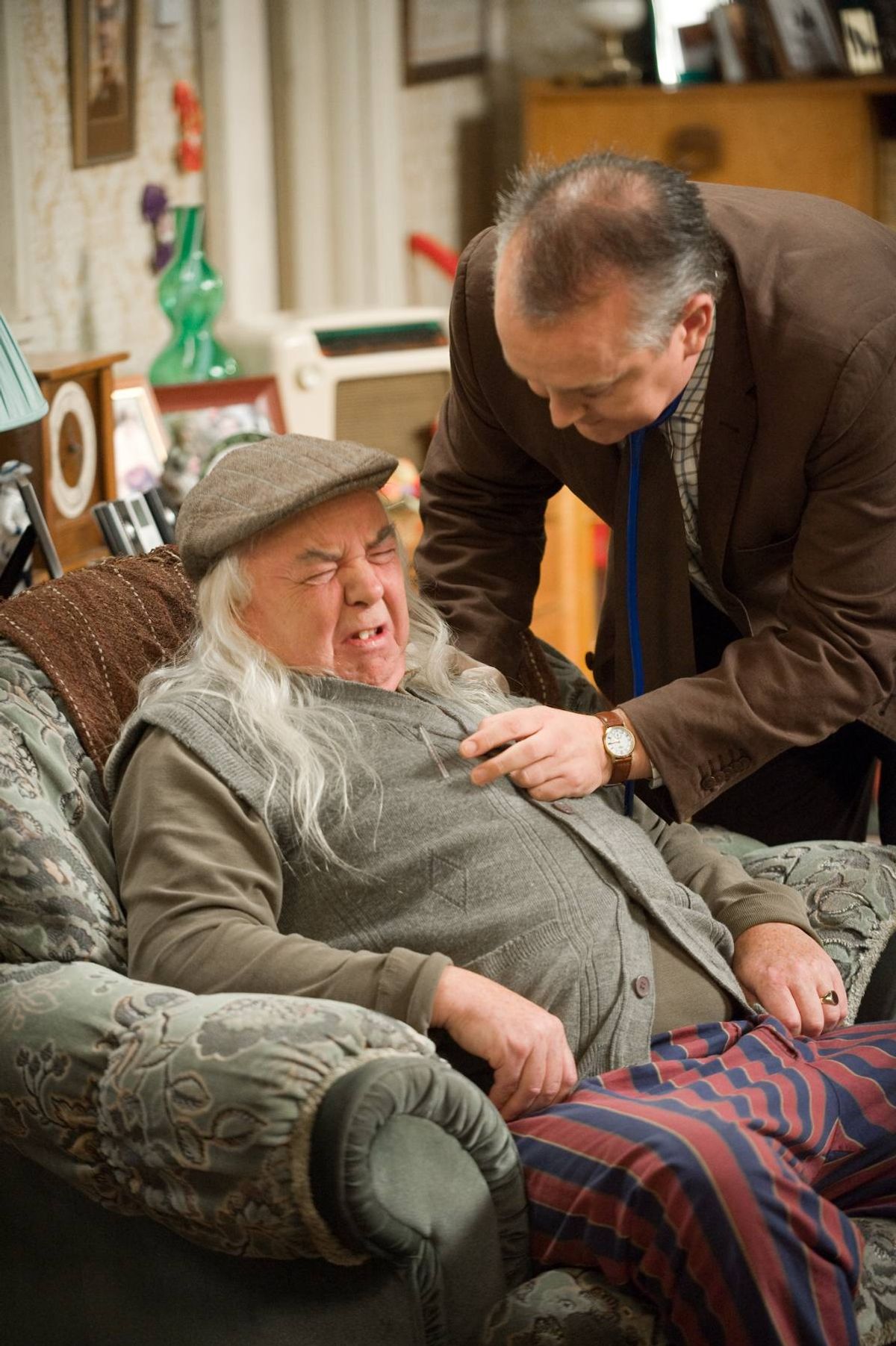An elderly individual, adorned with a cap and a long white beard, is seated on a couch, appearing visibly distressed and shouting. A middle-aged man in a suit and tie is leaning over, attentively observing the elderly person. In the background, various household items and decorative objects are visible.