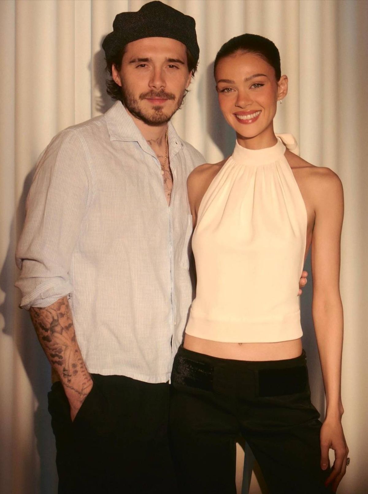 A man and a woman standing next to each other, posing for a photograph against a backdrop of white curtains. The man wears a white shirt and a hat, while the woman dons a sleeveless white dress.