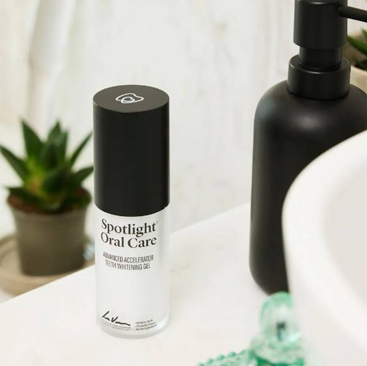 A cosmetic product in a black and white container is placed on a bathroom countertop. Adjacent to it is a small potted plant and a black bottle with a white lid. The countertop has a white surface and a wooden border.