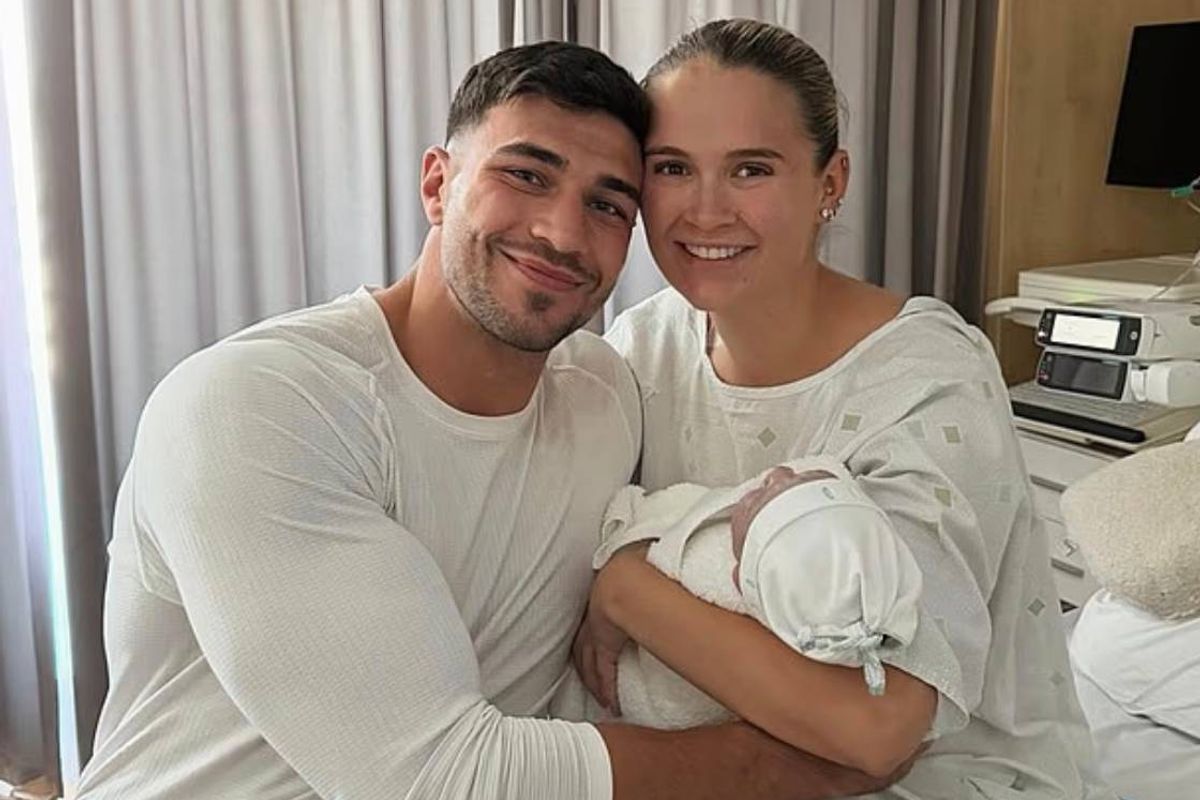 An adult male and an adult female are smiling and posing for a photograph while holding a newborn baby in a hospital room, with a window with white curtains in the background.