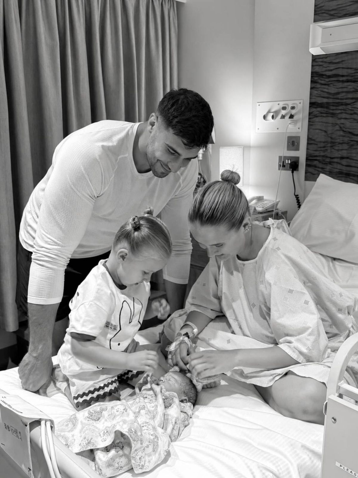A black-and-white photograph depicting a family moment in a hospital setting. An adult male stands beside a bed, gazing affectionately at a newborn baby lying in a hospital bed. A woman with a bun hairstyle, possibly the mother, is also present, looking towards the baby. A young child, presumably the couple's older child, stands nearby, looking curiously at the baby. The scene exudes warmth and familial bonding amidst the clinical environment.