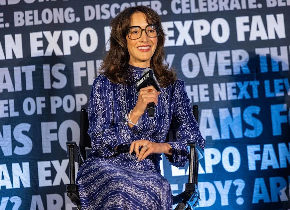 Jennifer Beals in a purple dress with long sleeves and a high neck on stage at FanExpo Chicago in 2025, sat on a chair and holding a microphone.