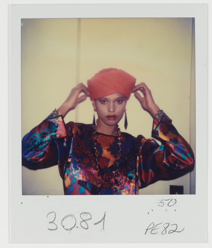 A person wearing a vibrant, multicolored patterned top and a bright orange turban stands against a plain background, adjusting the turban. The photo has handwritten numbers and notes at the bottom.