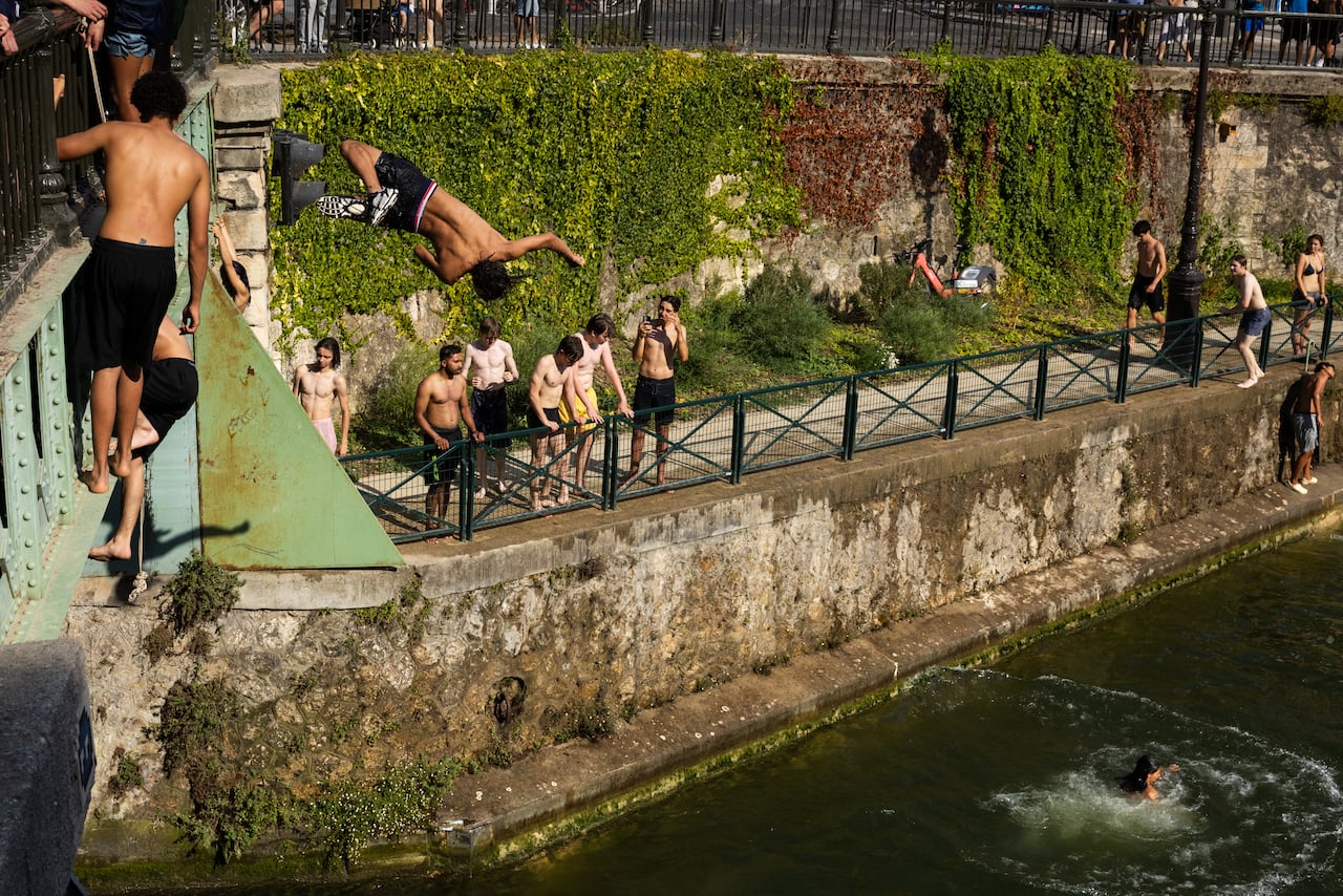 A person is shown twisting in the air in swim trunks above a natural body of water, as others stand on bridges above the water.