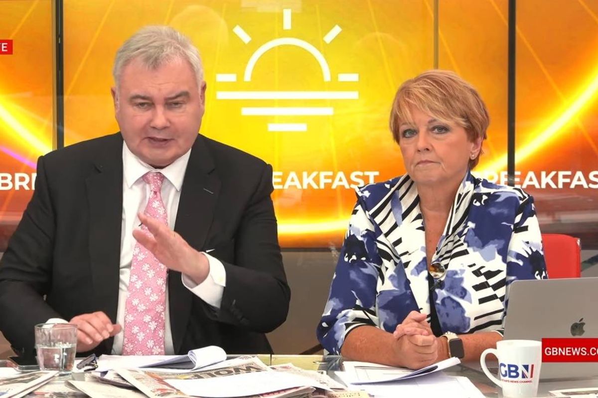 Two individuals are seated at a table in a studio setting, possibly engaged in an interview or discussion. The individual on the left wears a dark suit and a pink tie, while the person on the right is dressed in a blue and white patterned top. Behind them, a large screen displays a yellow backdrop with a sun graphic and some text. Various items such as papers, a laptop, and a cup are placed on the table in front of them.