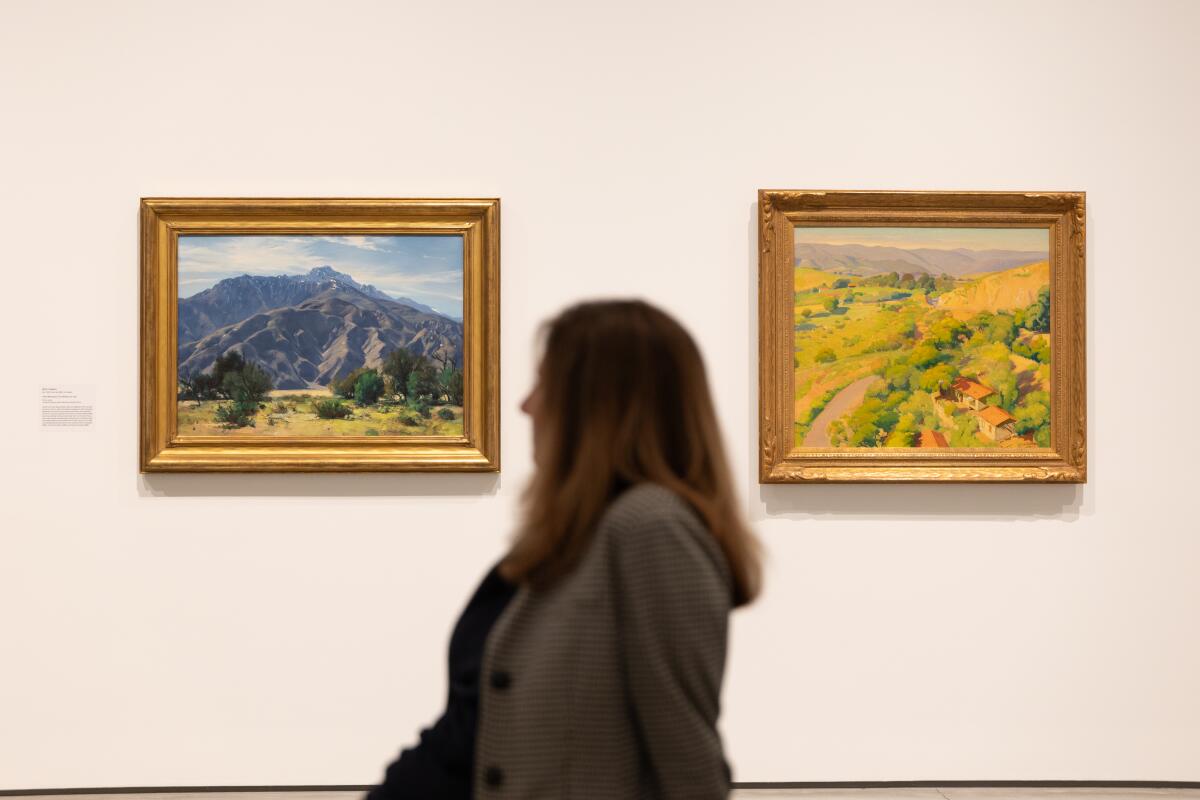 A woman stands between two landscape paintings, one of mountains and one of a yellow and green field.