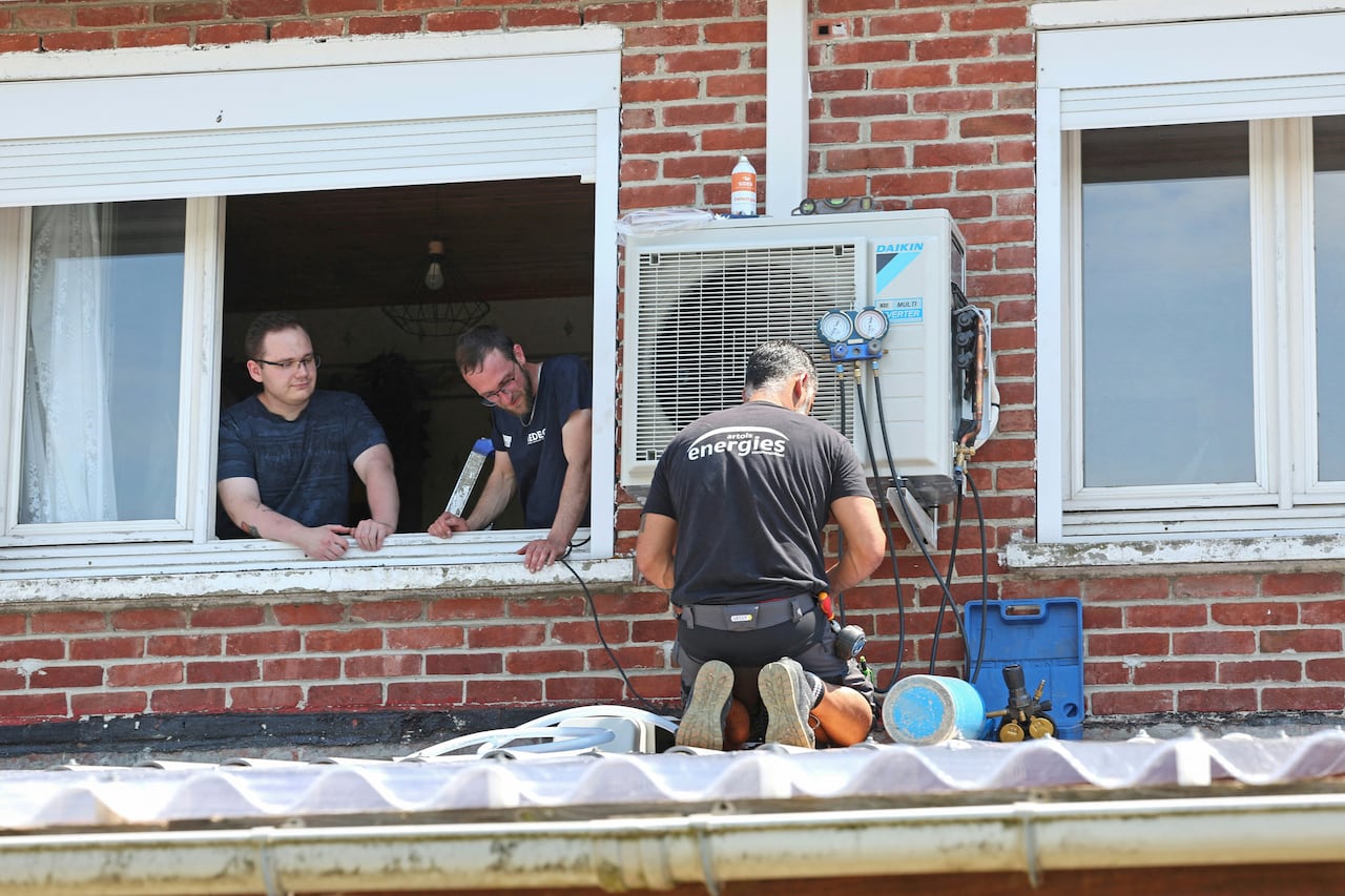 A man works on an air conditioning unit on a rooftop while two other people look on through a window.