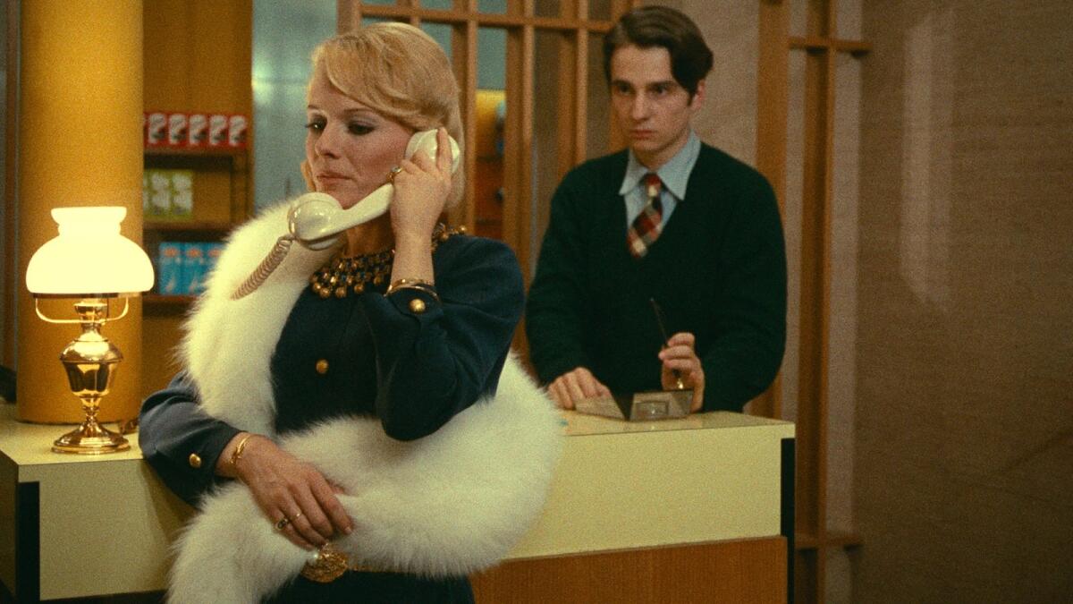 A glamorous woman makes a phone call while a man watches.