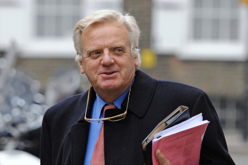 Man in a blue business shirt and navy coat carrying paperwork.