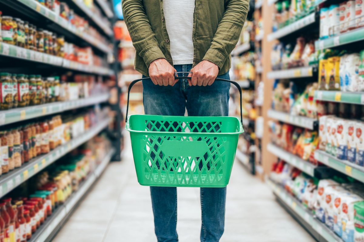A close-up of a man holding a green shopping basket while standing in the middle of a supermarket aisle. The shelves are stocked with colorful products, emphasizing the everyday activity of grocery shopping.
