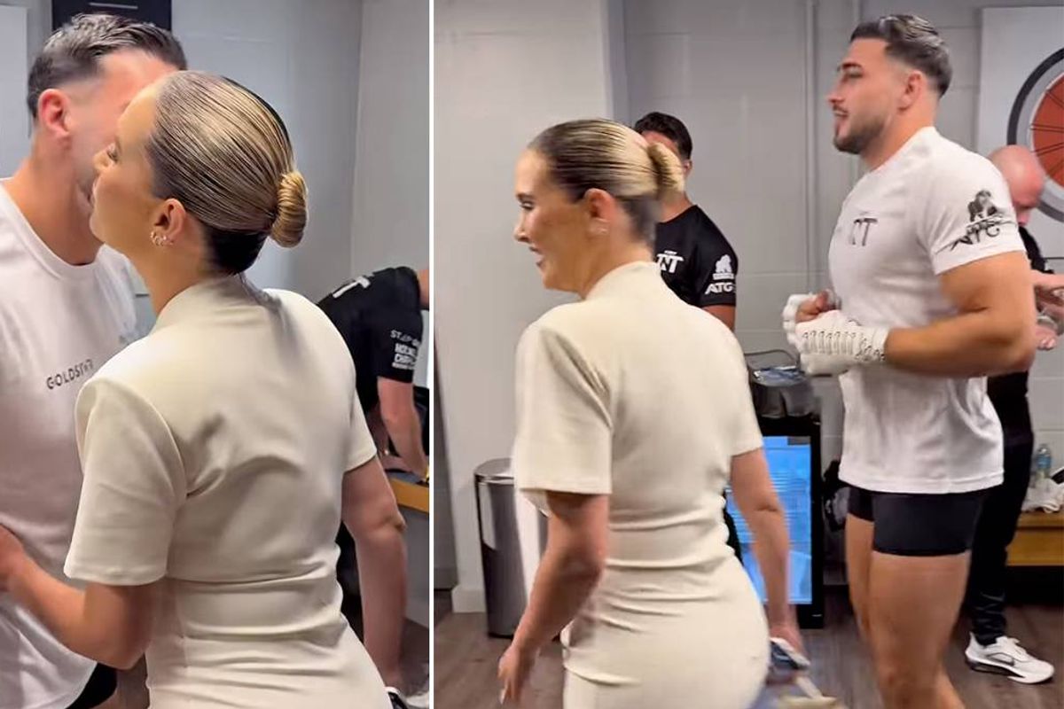 Two individuals, one wearing a white dress and the other in a white shirt and black shorts, engage in a conversation while being surrounded by other people in a room with white walls.