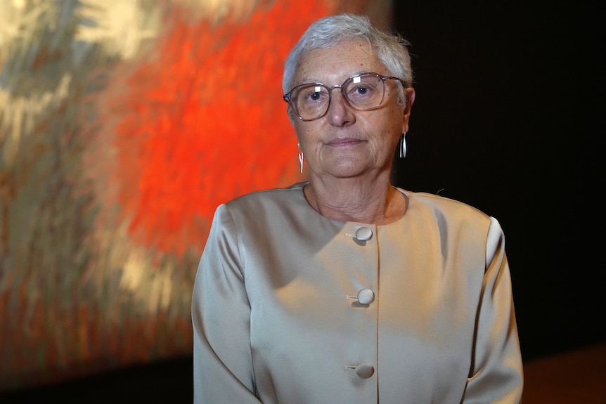 A woman with short grey hair and glasses stands in an art gallery.