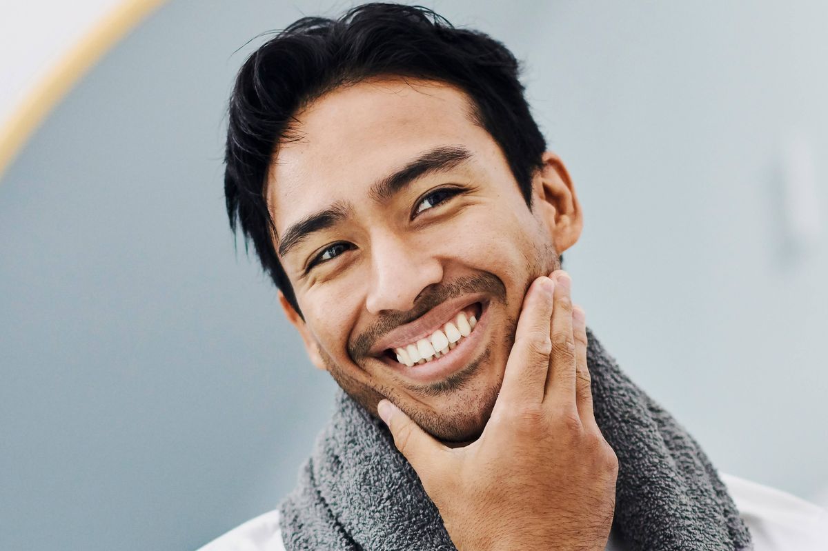 Man happy about face skin care product for healthy body and facial skincare for guys. Handsome young male looking in the mirror with toothy smile, touching his beard satisfied with his shaving cream