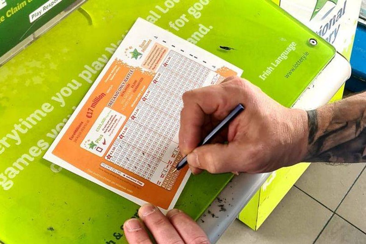 An individual with visible tattoos on their arm is signing a document with a pen. The document features a combination of orange and white sections and is placed on a green surface, possibly within an office setting.
