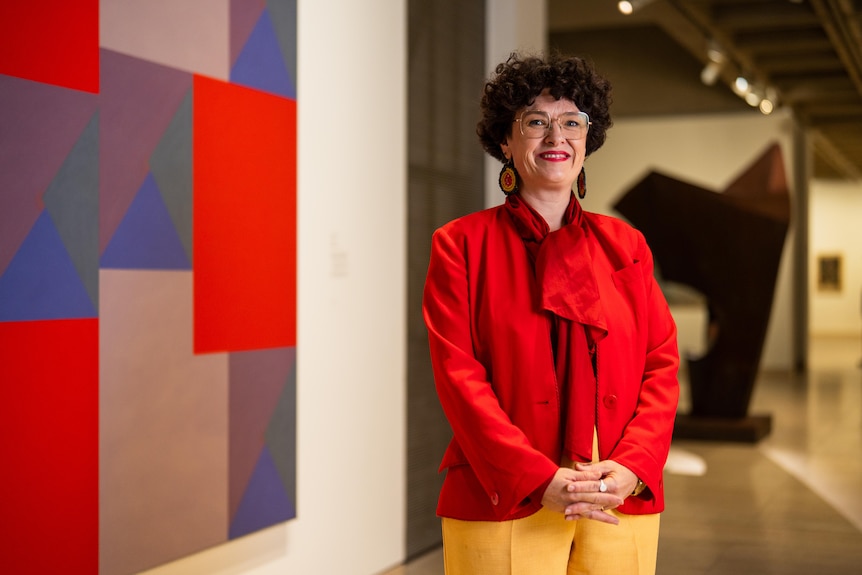 Bek Golsby-Smith with short curly brown hair, glasses, Aboriginal patterned earrings, a bright red jacket, smiling in a gallery.