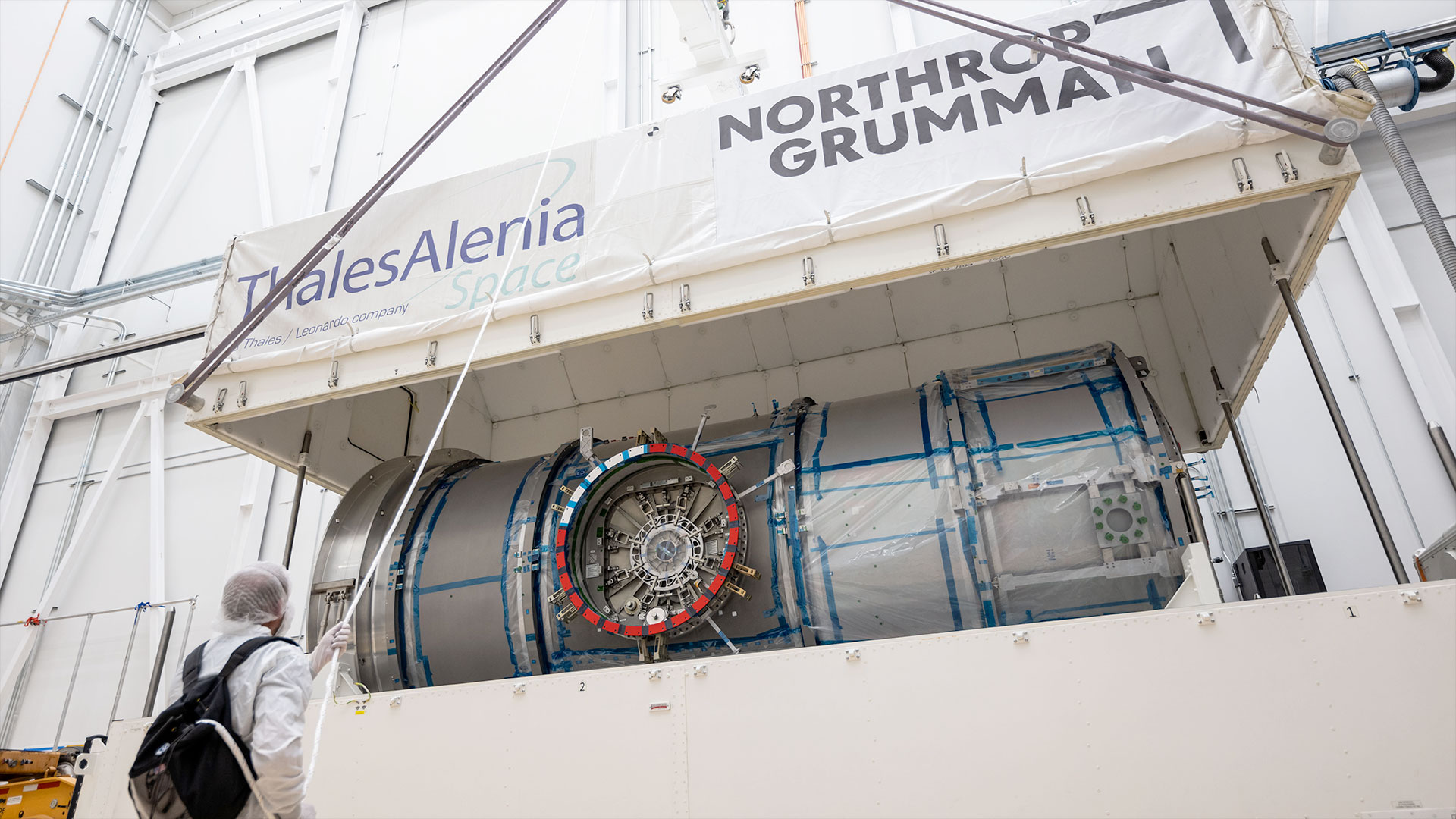 a white top half of a large shipping container is lifted by crane, exposing a metal cylindrical module inside.