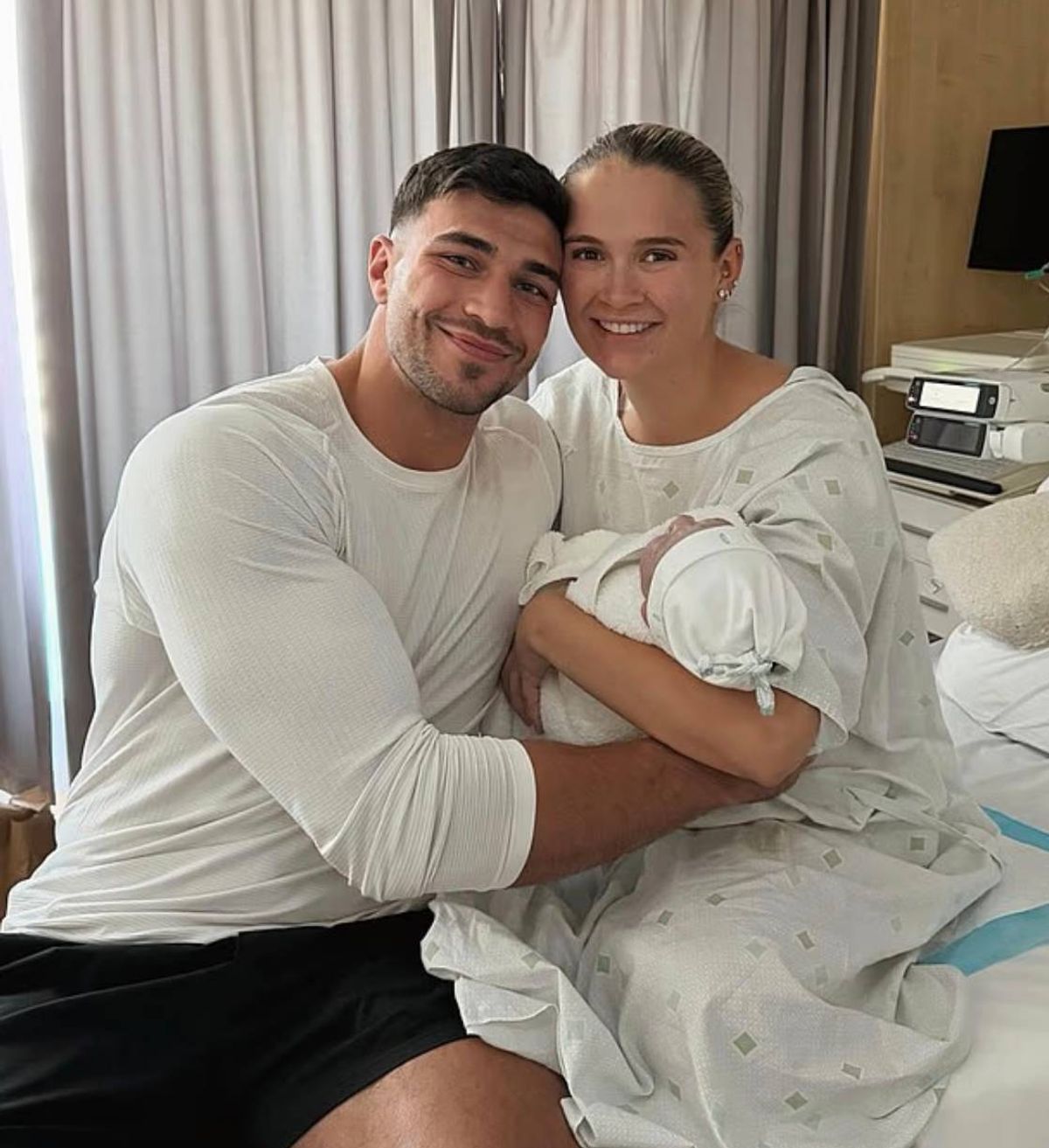 A smiling couple is holding a newborn baby in a hospital room, with a medical monitor and a lamp visible in the background.