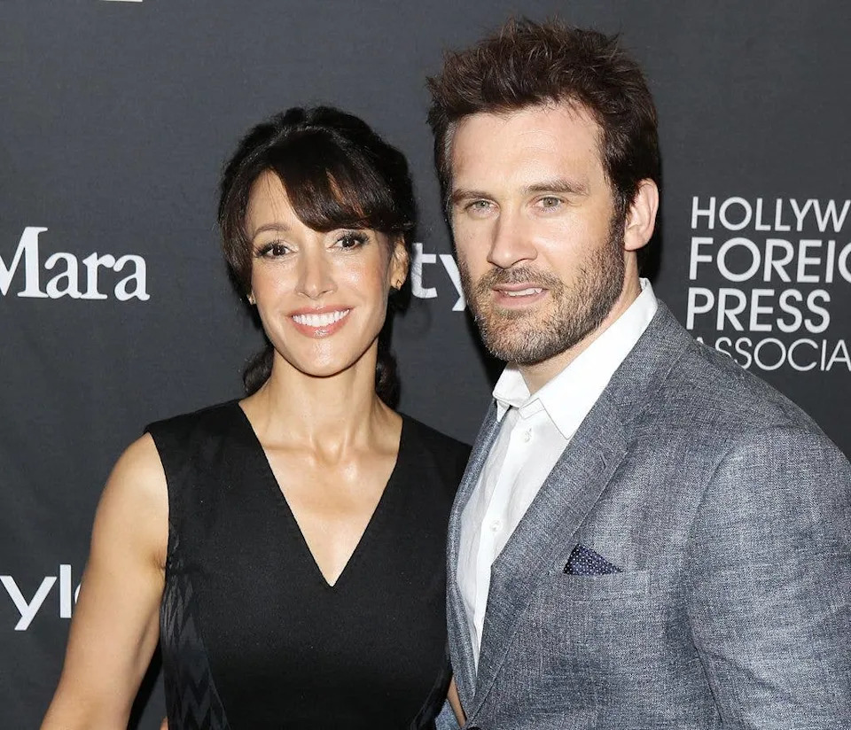 Jennifer Beals and Ken Dixon stand with an arm around one another. Jennifer wears a black v neck dress, Ken wears a metallic silver suit and white shirt.