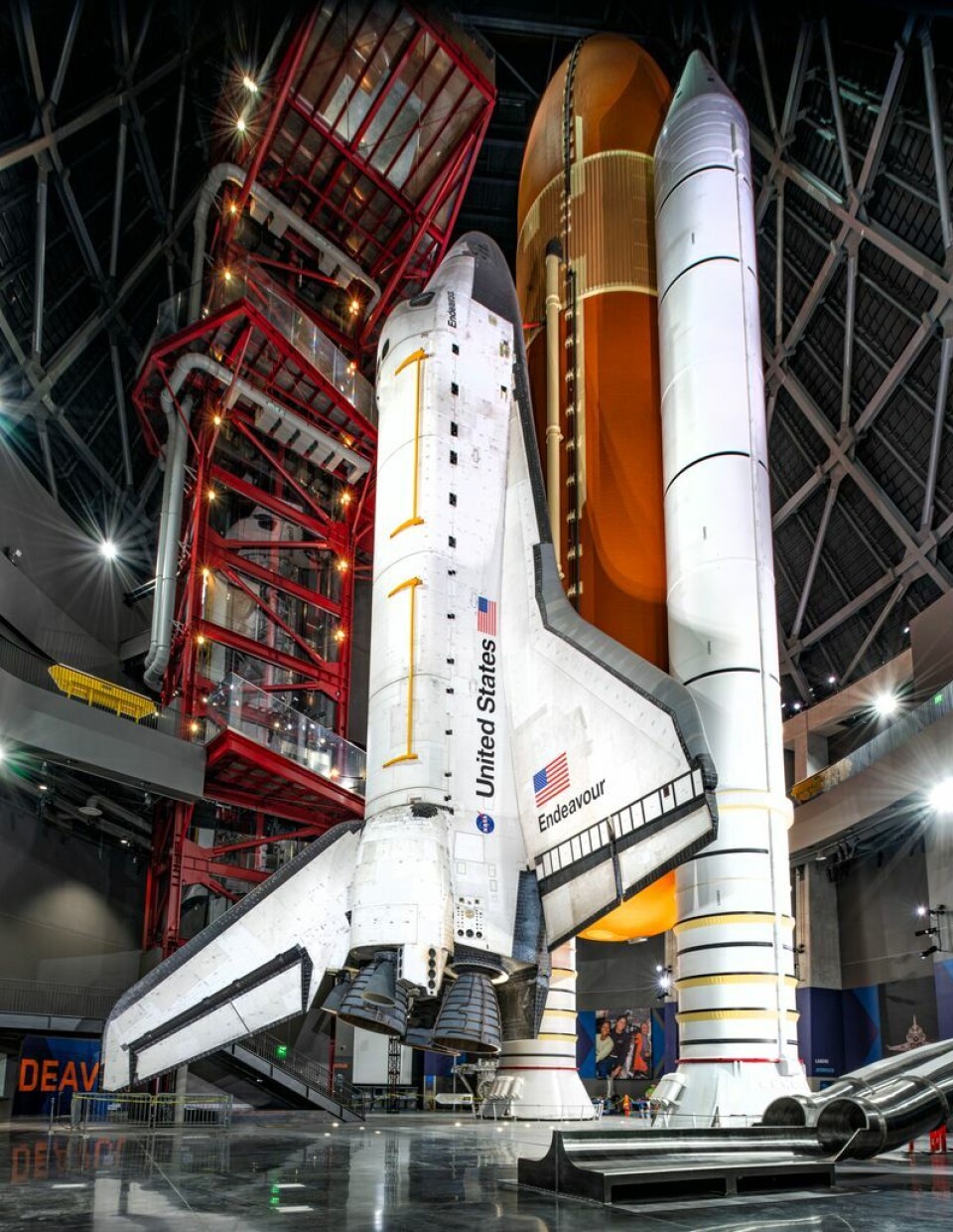 Space shuttle on display inside a hangar next to a booster and launch support structure.