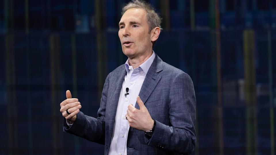 Andy Jassy wears a gray blazer over a light blue shirt and gestures with both hands while speaking on a darkened stage.