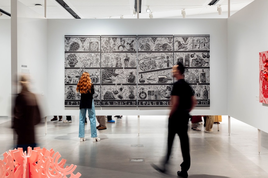Visitors in a gallery standing in front of a large black-and-white vinylcut print etched with images and patterns.