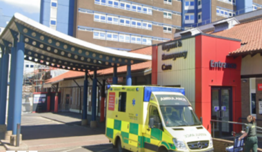 The exterior of the University Hospital of North Tees. The brick building has signs on it that read: "Urgent & Emergency Care". There is an ambulance parked out front and a paramedic pushing a wheelchair.