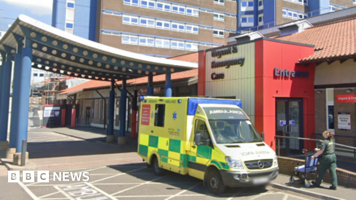 The exterior of the University Hospital of North Tees. The brick building has signs on it that read: "Urgent & Emergency Care". There is an ambulance parked out front and a paramedic pushing a wheelchair.