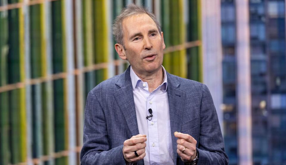 Andy Jassy wears a gray blazer over a light blue open-collar shirt and gestures with both hands while speaking, with a colorful striped backdrop behind him.