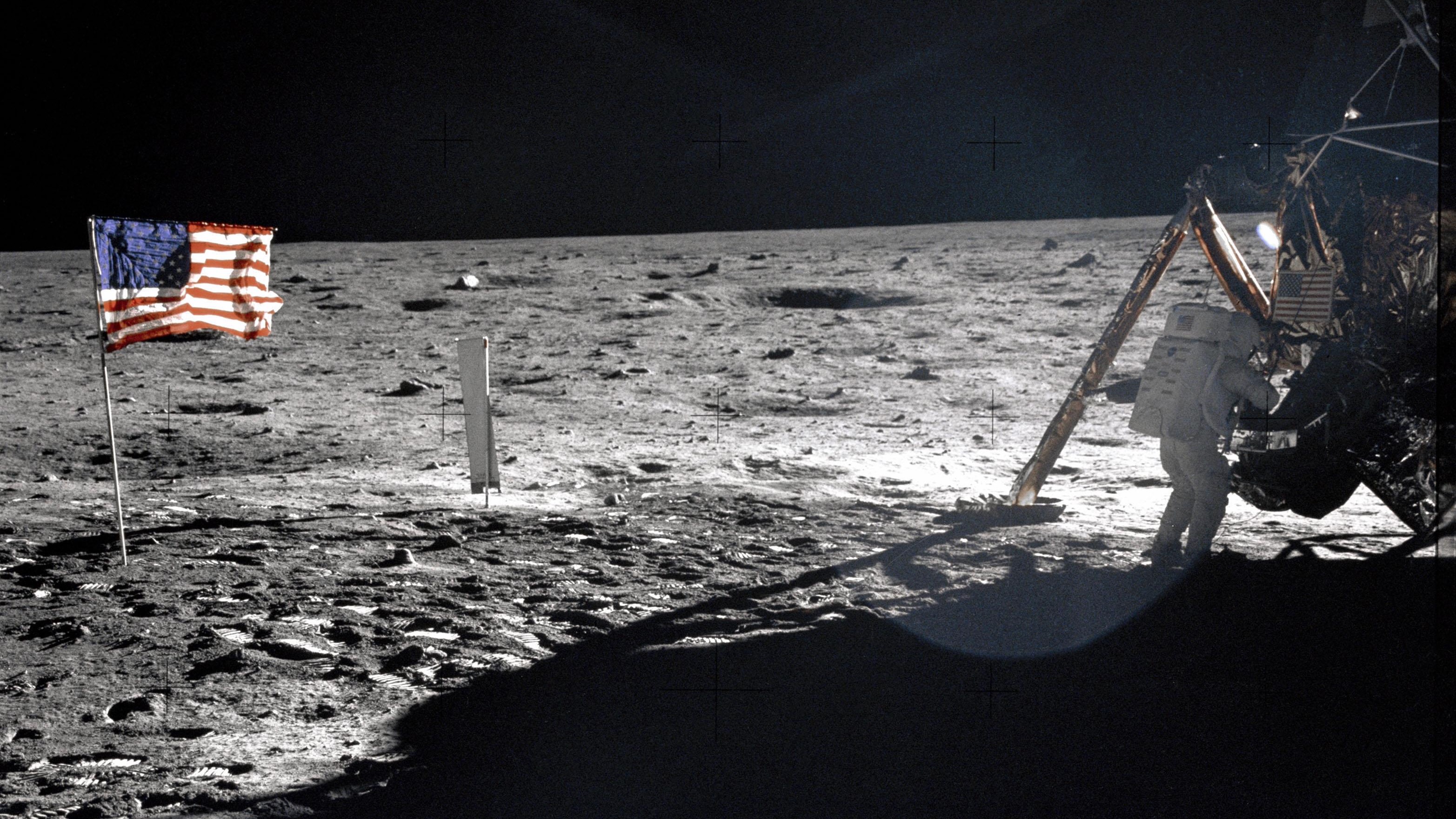 an astronaut in a white spacesuit descends from a gold-foil-wrapped spacecraft onto the gray lunar surface, with an american flag planted in the dirt nearby
