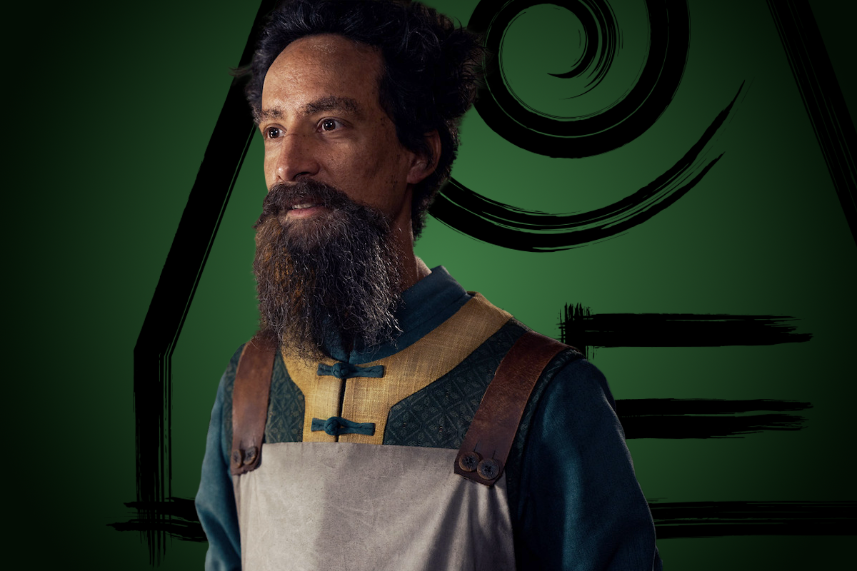 Danny Pudi as Sai (The Mechanist) in fantasy attire with leather straps stands before a dark green background featuring abstract spiral and line designs, suggesting a mystical or fictional environment.
