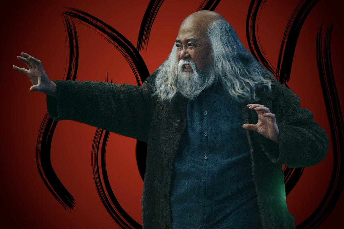 Momona Tamada as Uncle Iroh with long gray hair and beard, wearing a dark fur coat, stands with arms raised against a red background featuring black flame-like patterns, appearing intense and focused as if performing or casting something.