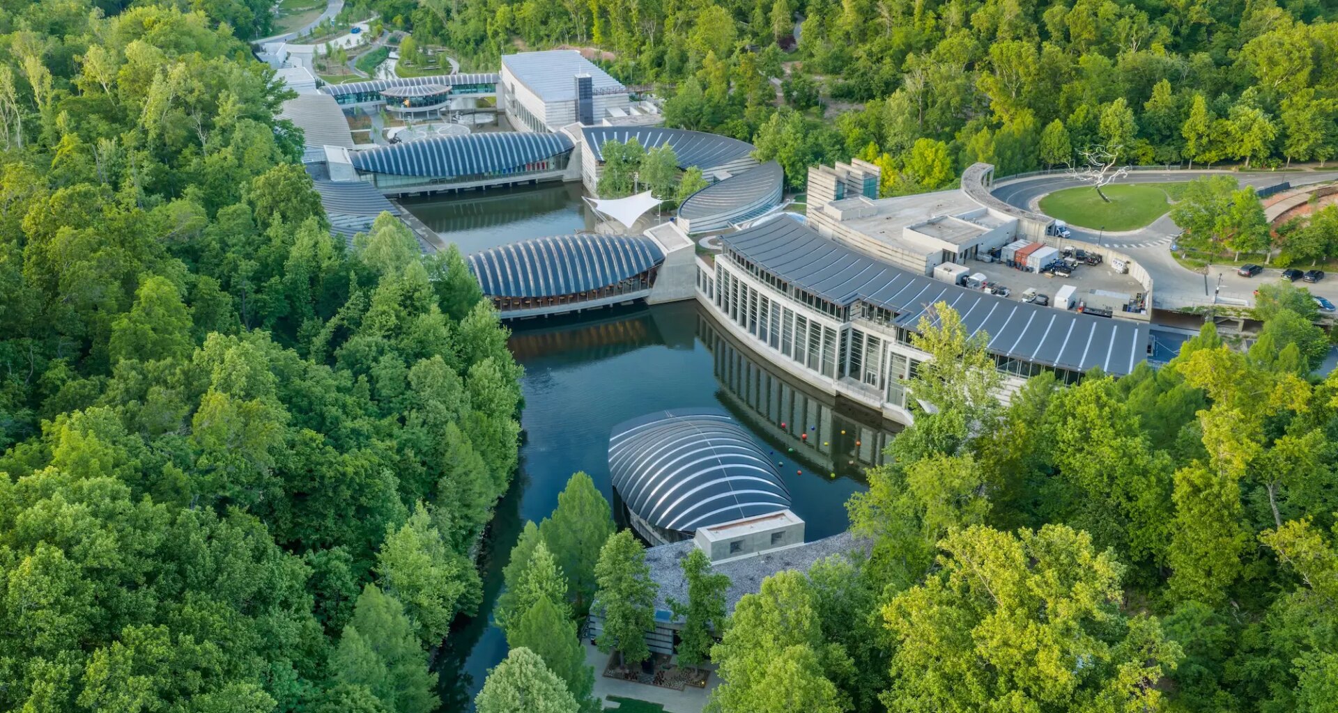 Aerial view of modern architecture museum surrounded by lush green forest and water in a scenic landscape.