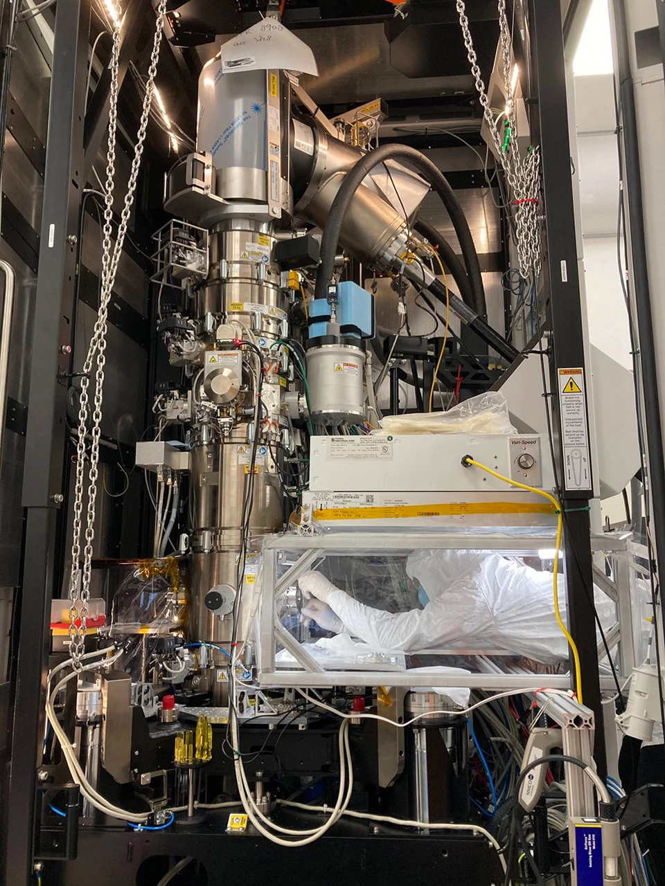 A person in a cleanroom suit works hunched under a HEPA filter inside the chassis of an electron microscope, which features lots of hardware.