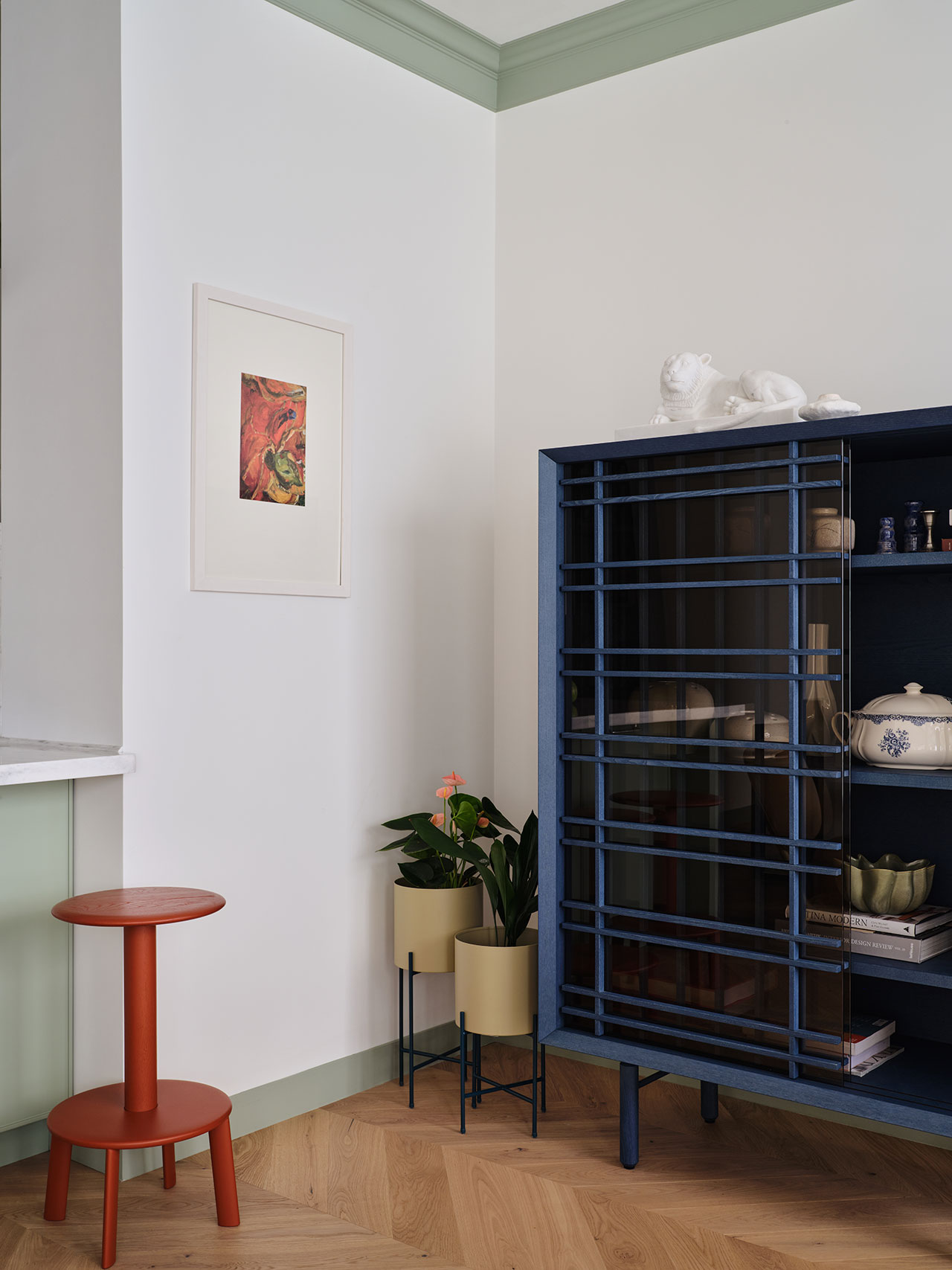 A corner interior with a blue cabinet, a red stool, two potted plants, and framed art on a white wall with green accents and herringbone wood flooring.