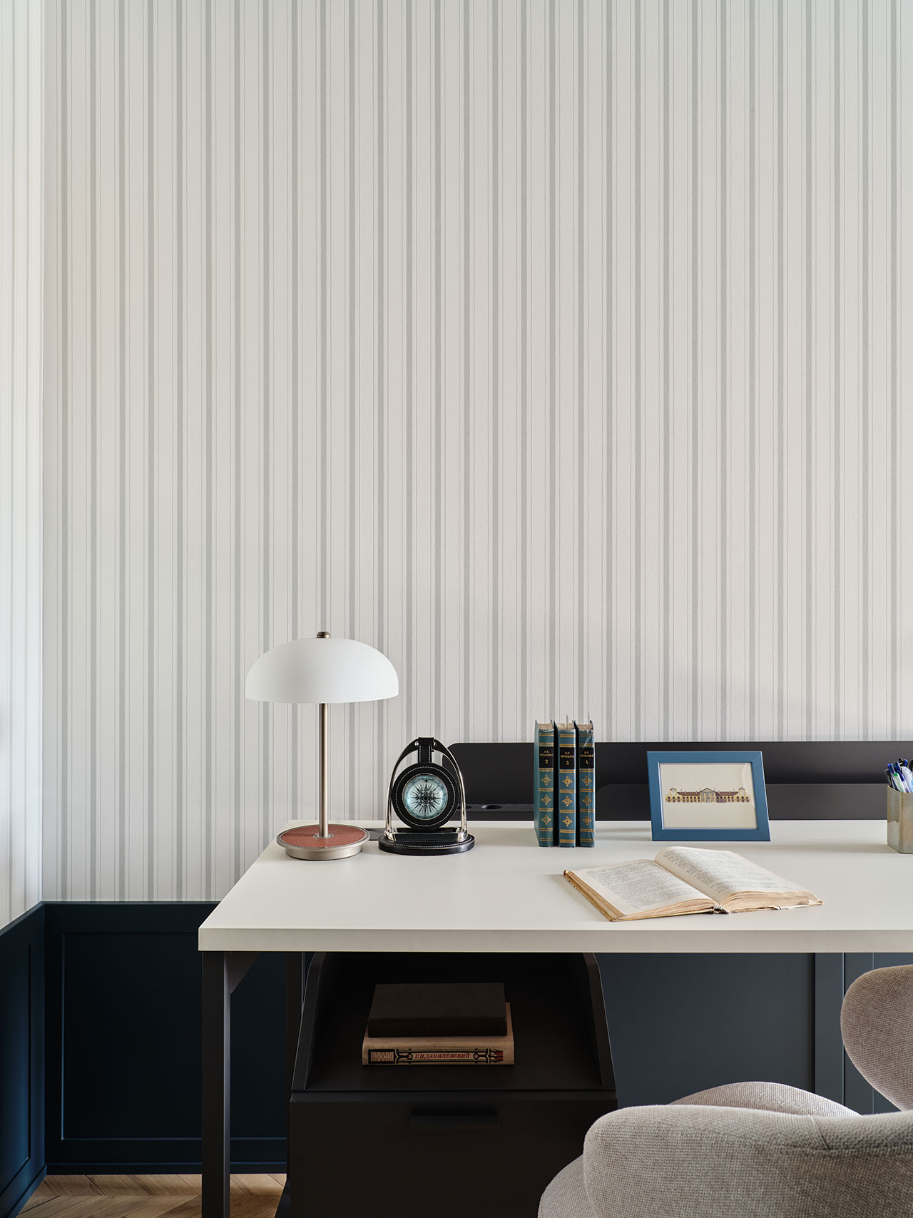 A modern desk with an open book, a lamp, a clock, stacked books, a framed picture, and a pencil holder against a striped wall.