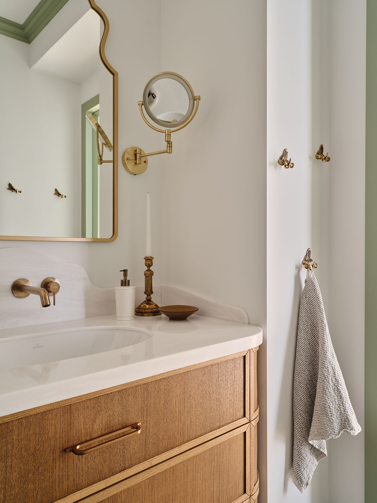 Modern bathroom vanity with a wood cabinet, white countertop, brass fixtures, wall-mounted mirror, candle holder, soap dispenser, and a towel hanging on a brass hook.