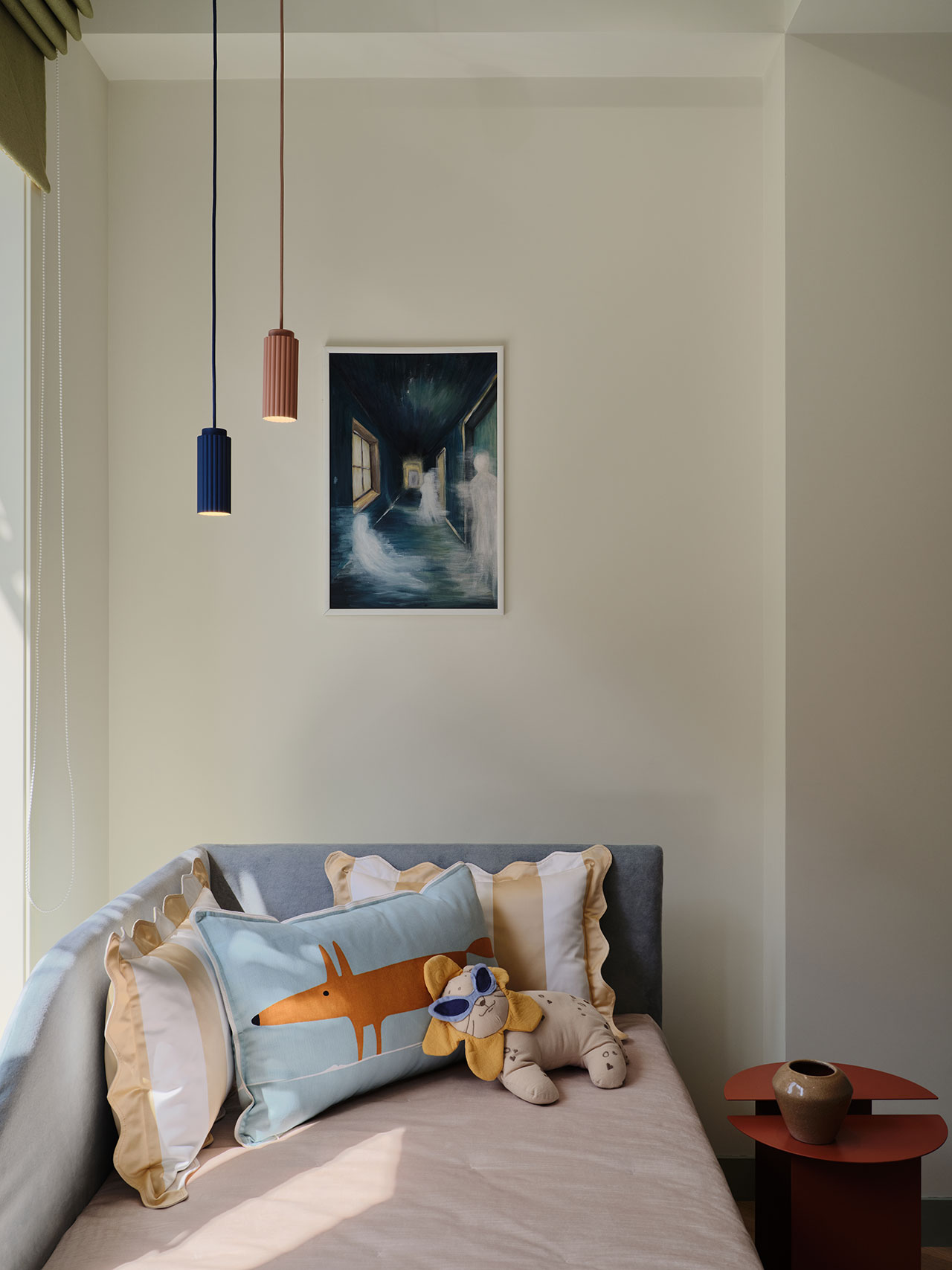 A small sofa bed with decorative pillows and a plush toy sits beneath a framed painting; pendant lights hang above, and a side table is nearby.