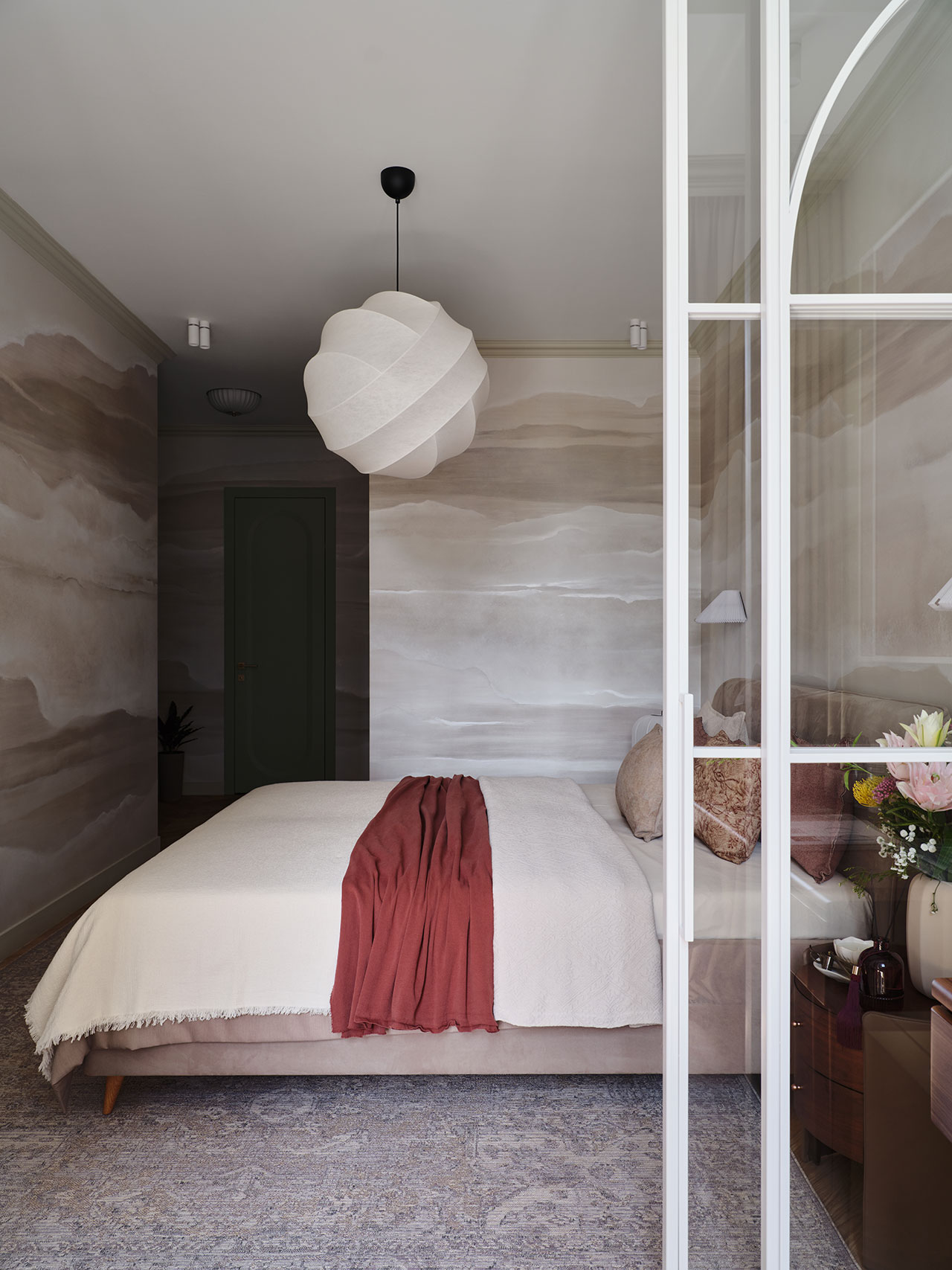 A modern bedroom with a beige bed, a white and red blanket, muted abstract wall art, a round white pendant light, and a glass door partially open. Flowers sit on the bedside table.