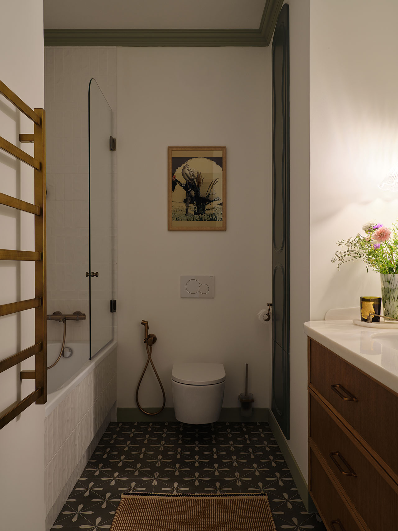 Modern bathroom with patterned tile floor, wall-mounted toilet, glass-panel bathtub, wooden vanity, brass towel rack, and framed artwork above the toilet. Flowers sit on the sink.