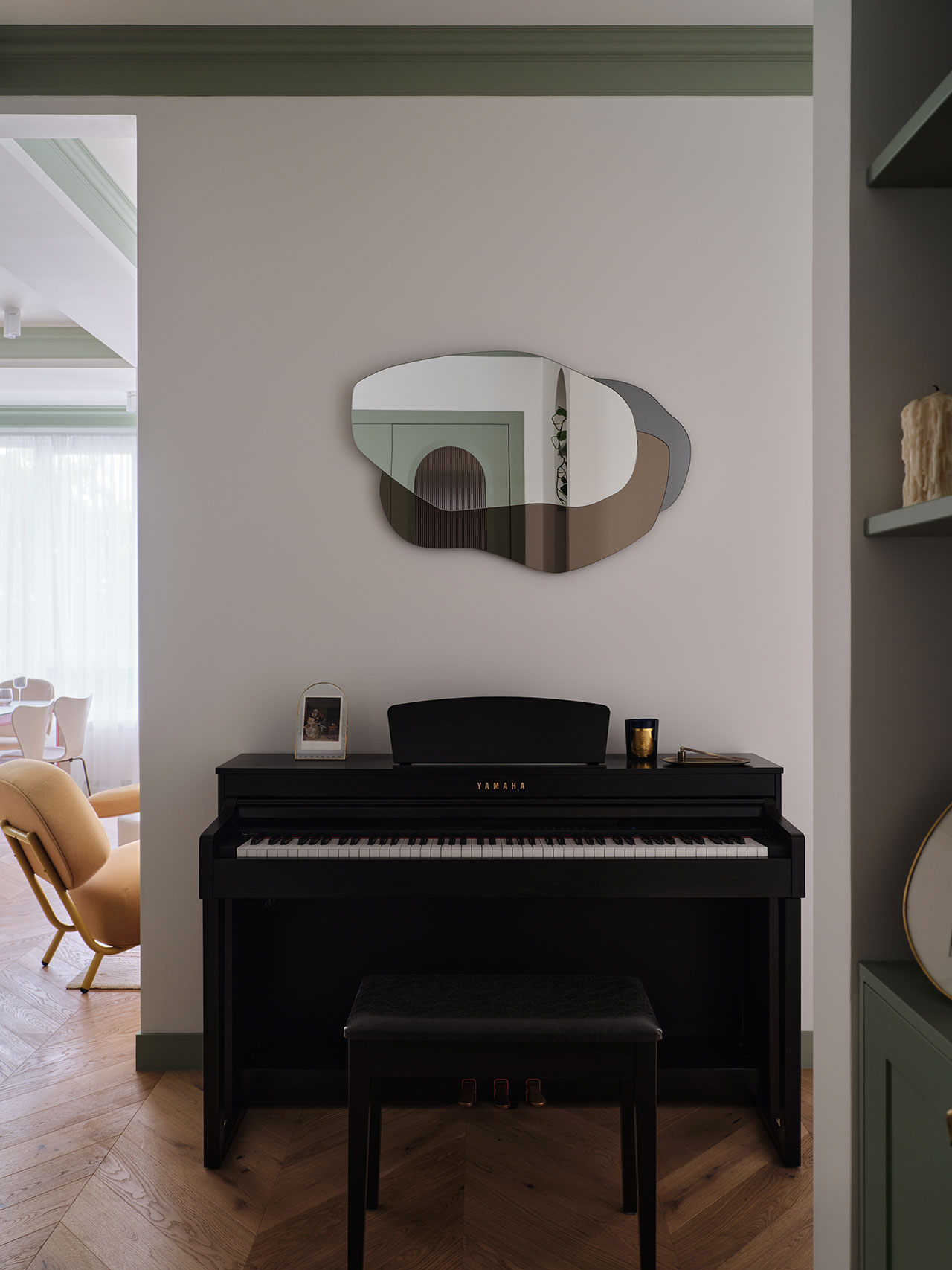 A black Yamaha piano with a bench sits against a white wall, topped with a decorative mirror, photo frame, and candle; a yellow chair is visible in the adjacent room.