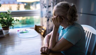 Older woman in blue shirt sitting at kitchen table, gazing pensively out window