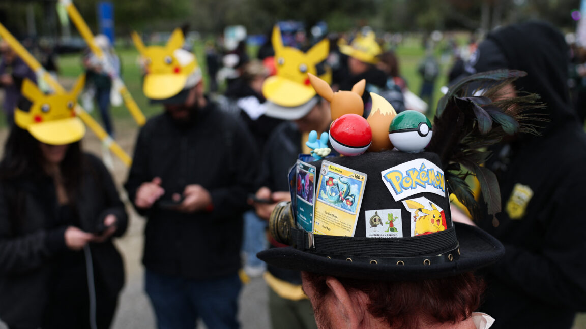 A player wearing a hat decorated with Pokemon characters and trading cards plays Pokemon GO on a smartphone during the in-person Pokemon GO Tour: Kalos Los Angeles 2026 event at the Rose Bowl Stadium in Pasadena, California, on February 20, 2026.