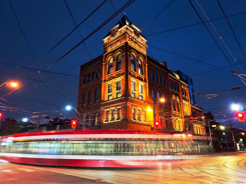 A historic brick building illuminated at night, with a red and white streetcar in motion creating light trails, surrounded by traffic lights and overhead wires.