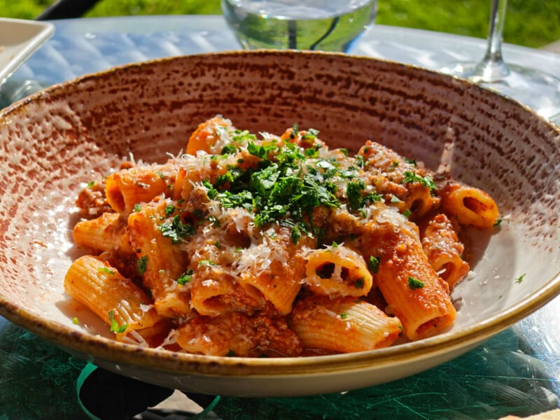 A bowl of rigatoni pasta topped with meat sauce, grated cheese, and chopped parsley, served on a speckled ceramic plate, with sunlight shining on the dish.