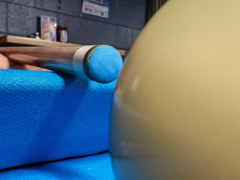 Close-up of a blue-tipped pool cue aimed at a cue ball on a blue billiards table, with part of a wall and other objects blurred in the background.