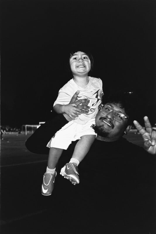 A smiling adult lifts a young child onto their shoulder at night. The child grins and wears sporty shoes, while the adult makes a peace sign and smiles, both looking happy outdoors.