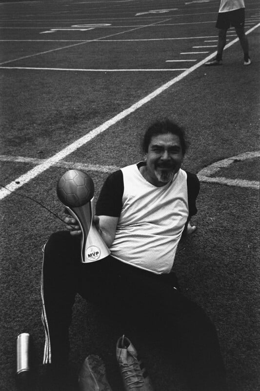 A man sits on a sports field, smiling and holding a trophy labeled "MVP" and a small soccer ball. He wears a light-colored shirt with dark sleeves and athletic pants. Another person stands in the background.