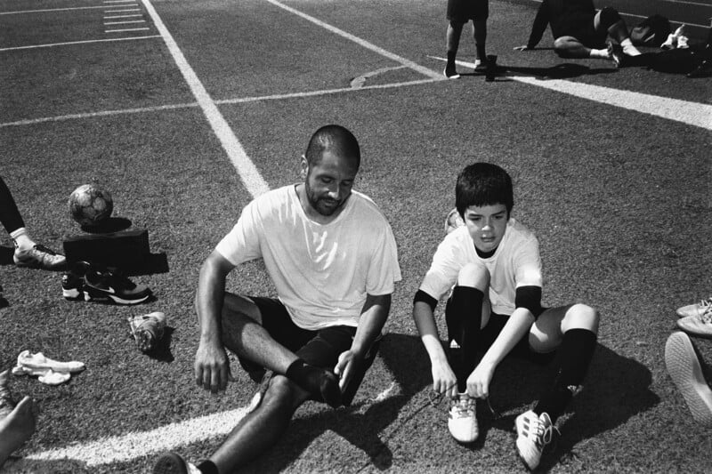 A man and a boy sit on a sports field, wearing athletic gear. The man smiles while the boy ties his shoe. Soccer shoes, a ball, and other players are visible around them on the grassy field.