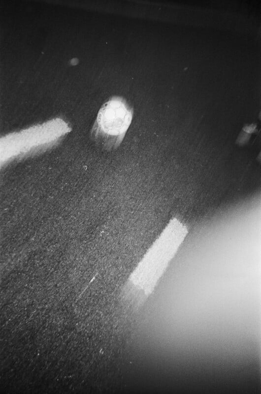 A blurry black and white photo shows a soccer ball in motion on a street at night, with two white road markings visible on the asphalt.